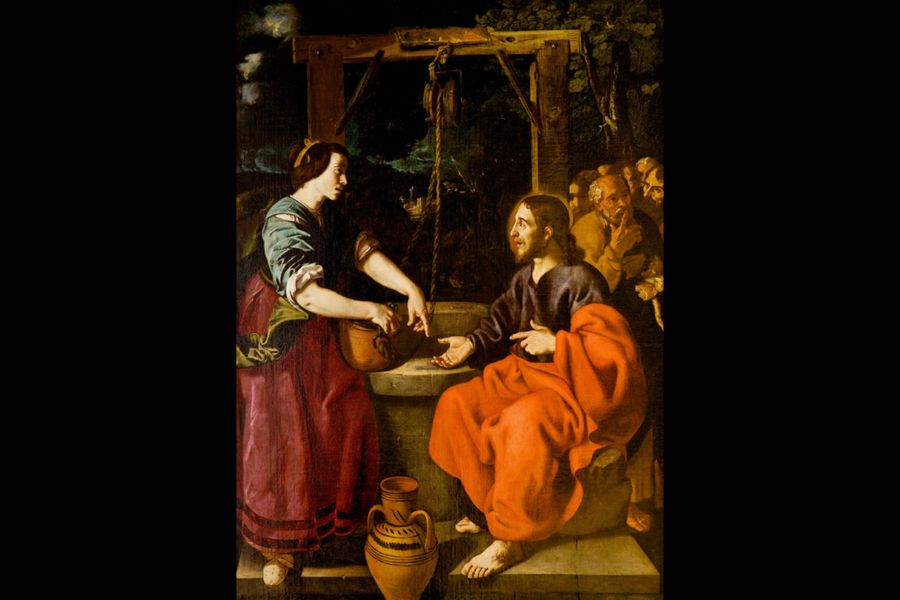 A painting in a Baroque style showing a biblical scene at a stone well at night: on the left, a woman in a red skirt, green sash, and teal blouse draws water from a bucket, while on the right a seated, barefoot robed man in a deep blue tunic and bright orange-red cloak gestures toward her, with clay water jugs at their feet and a group of onlookers gathered in the dark background beneath a wooden well structure and foliage.