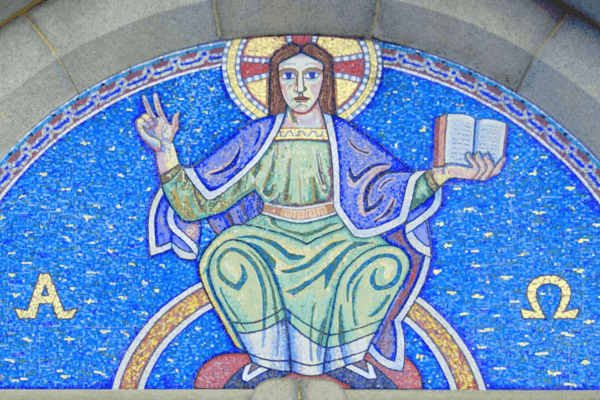 Mosaic artwork of a seated haloed religious figure with the face blurred, raising one hand in blessing and holding an open book, set against a bright blue background with alpha and omega symbols on either side.