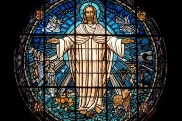 A circular stained glass window depicting Jesus Christ standing in the center with arms outstretched, dressed in white robes with a golden halo behind his head. He is surrounded by blue glass radiating outward in sunburst-like rays. Surrounding figures include a lamb at his feet, doves in flight, an angel to his left, and various Christian symbols including a cross and a chalice. The window is set against a black background