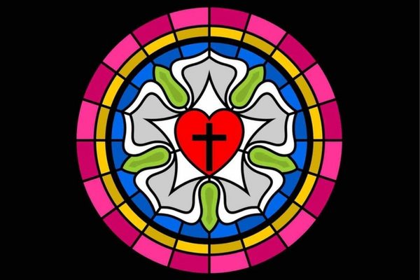 Stained-glass Luther Rose: a red heart with a black cross at the centre of a white five-petalled rose with grey shading and green sepals, set in a blue circle ringed by gold and bright pink glass panels, on a black background.